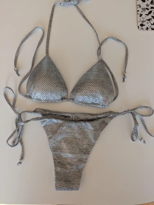 Silvrig bikini Calzedonia XS  - Silverskimrande bikini med småmönstrad yta och knyt med pärlor i både topp och trosa. Triangelformad överdel och klassisk bikiniunderdel med knyt i sidorna. Märke Calzedonia, stlk XS. Mycket fint skick.