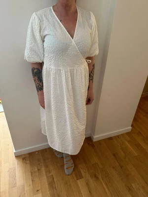 Vit omlottklänning med struktur - Säljer en vit omlottklänning med struktur och knyt i sidan. Klänningen har v-ringning, puffiga korta ärmar och en luftig passform. Perfekt för sommarens varma dagar och har en stilren, enkel look.