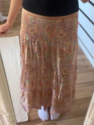 Mönstrad långkjol i korall - Luftig långkjol med bohemiskt mönster i pastelliga toner av rosa, orange, beige och grönt. Kjolen har smock i midjan för en bekväm passform och ett flowy fall. Perfekt för varma dagar.
