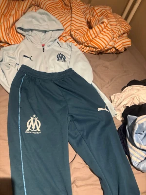 Marseille träningsset från Puma - Säljer ett Marseille träningsset från Puma med ljusblå huvtröja och mörkblå byxor. Setet har klubbens logga och Puma-logga på både tröja och byxor. Huvtröjan har dragkedja och fickor, byxorna har resår i midjan och ljusblå detalj längs sidan.