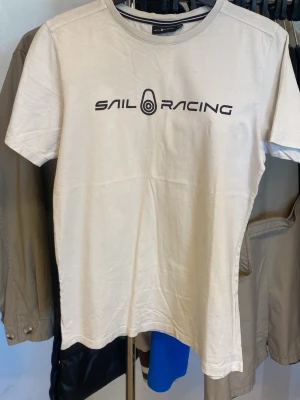 Vit Sail Racing t-shirt i bomull - Vit t-shirt från Sail Racing med svart logotyptryck på bröstet och en liten logga i nacken. Klassisk passform och rund halsringning. Tillverkad i mjuk bomull som känns skön mot huden. Perfekt för en clean och sportig stil.