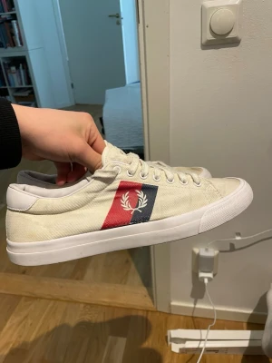 Vita Fred Perry sneakers med logga - Snygga vita sneakers från Fred Perry med klassisk logga på sidan i rött och blått. Skorna har lågt skaft, vita snören och är tillverkade i canvasmaterial med gummisula. Perfekta för en stilren och sportig look.
