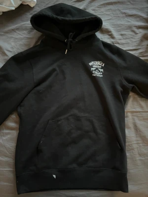 Svart hoodie  Zach Bryan - Svart hoodie med tryck på bröstet där det står Ontario CA och en bil, samt stort tryck på ryggen med Zach Bryan och låtlista. Klassisk känguruficka framtill och dragsko i huvan. Perfekt för dig som gillar musikmerch och streetwear. Den har en liten färg fläck på framsidan.