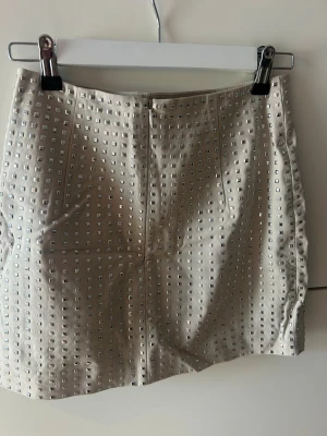 Silverfärgad kortkjol med nitar - Säljer en snygg kortkjol i silver med fyrkantiga nitar över hela kjolen. Kjolen har en rak passform och är tillverkad i ett glansigt material som ger en cool look. Perfekt för dig som vill sticka ut med en edgy stil. Storlek 36, S. 