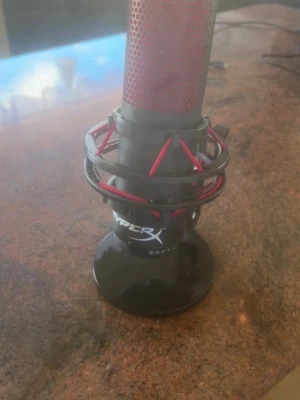 HyperX röd och svart mikrofon - Säljer en HyperX mikrofon i svart och rött med metallgaller och stötdämpande upphängning. Mikrofonen har en rund bas och tydlig HyperX-logga. Perfekt för streaming, gaming eller poddande.