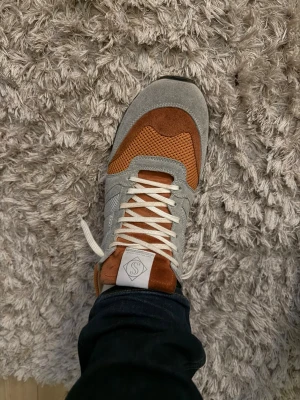 Grå och orange sneakers från Sneakersnstuff - Snygga sneakers i grått och orange från Sneakersnstuff. Ovandelen är i mocka och mesh med vita skosnören och en vit logga på plösen. Skorna har en klassisk låg silhuett och passar perfekt till jeans eller streetwear.