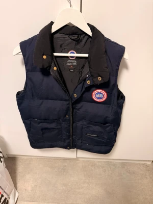 Mörkblå dunväst från Canada Goose - Snygg mörkblå dunväst från Canada Goose med klassisk logga på bröstet och två stora fickor framtill. Västen har hög krage, dragkedja och tryckknappar. Perfekt för lager-på-lager och håller dig varm under kyliga dagar.