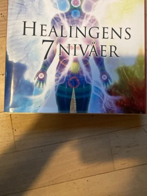 Healingens 7 nivåer bok - En färgstark bok med titeln 'Healingens 7 nivåer'. Omslaget har en andlig illustration med chakran och energiflöden i blått, lila och gult. Boken är inbunden och har ett konstnärligt, spirituellt tema. Perfekt för dig som är intresserad av healing och personlig utveckling.