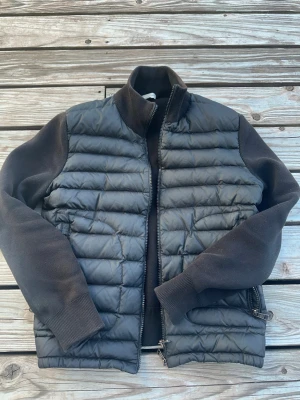 Svart dunväst från Moncler - Snygg svart dunväst från Moncler med quiltad framsida och baksida samt stickade svarta ärmar och muddar. Västen har hög krage och dragkedja framtill. Perfekt för lager-på-lager och streetstyle.