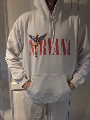 Vit Nirvana hoodie med tryck - Säljer en vit hoodie med stor huva och magficka. På framsidan står det 'Nirvana' i rött med en ikonisk ängelgrafik, och på baksidan finns samma ängelmotiv och text. Perfekt för dig som gillar bandmerch och vill ha en skön, oversized stil.