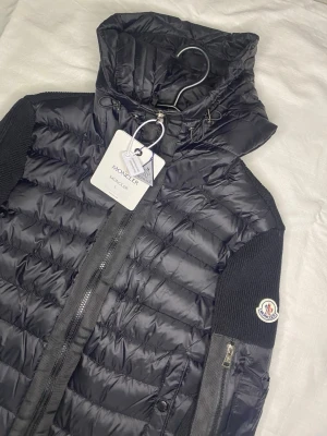 Svart pufferjacka från Moncler - Svart pufferjacka från Moncler med stickade ärmar och huva. Jackan har dragkedja framtill, två fickor med knappar och en extra ficka med dragkedja på ärmen. Moncler-logga på vänster ärm. Materialmix av dun och stickat ger en modern look.