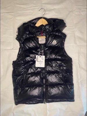 Svart dunväst från Moncler med huva - Snygg svart dunväst från Moncler med glansig finish, avtagbar huva och två dragkedjefickor. Klassisk Moncler-logga på bröstet och dragkedja framtill. Perfekt för lager-på-lager och streetstyle.