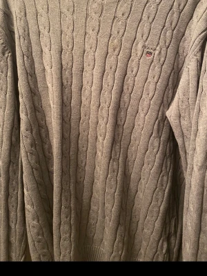 Beige stickad tröja Gant - Kabelstickad beige tröja från Gant med rund halsringning och diskret logga broderad på bröstet. Tröjan har långa ärmar och ribbade muddar vid ärmslut och nederkant. Perfekt för lager på lager och klassisk stil.