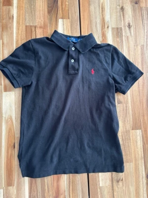 Svart pikétröja från Polo Ralph Lauren - Klassisk svart pikétröja från Polo Ralph Lauren med korta ärmar och krage. Tröjan har två knappar framtill och den ikoniska röda logotypen broderad på bröstet. Perfekt för en stilren och avslappnad look.