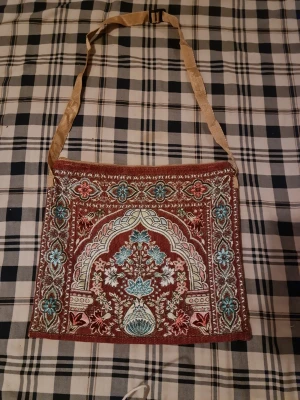 Broderad axelväska med orientaliskt mönster - Unik axelväska i tyg med broderat orientaliskt mönster i turkos, vitt och rött på brun bakgrund. Väskan har en beige axelrem och fyrkantig form. Perfekt för dig som gillar bohemisk och färgstark stil.