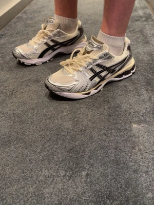Asics Gel-Kayano 14  - Snygga Asics Gel-Kayano 14 sneakers i vitt och silver med svarta detaljer. Skorna har en sportig mesh- och syntetöverdel, klassisk Asics-logga på sidan och chunky sula med gel-dämpning. Skorna är i storlek 44 men passar dig med 43