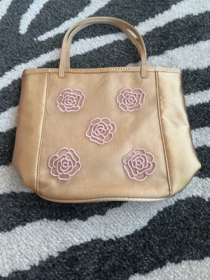 Beige handväska med rosa blommor - Snygg beige handväska med två handtag och broderade rosa blommor på framsidan. 18 cm lång och 24 cm bred