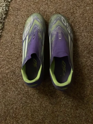 Lila Adidas F50 fotbollsskor - Säljer ett par lila Adidas F50 fotbollsskor med neongula detaljer och vita ränder. Skorna har en strömlinjeformad design utan snörning och är tillverkade i syntetmaterial för lätt vikt och bra bollkontroll. Perfekta för dig som vill sticka ut på planen.