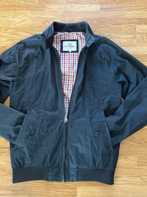 Svart jacka Ben Sherman - Stilren svart vindjacka från Ben Sherman med dragkedja framtill, två snedställda fickor med knapp och ribbade muddar. Fodret har ett klassiskt rutigt mönster i rött och vitt. Perfekt för blåsiga dagar och enkel att matcha med olika outfits.