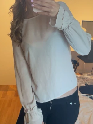 Beiget blus  - Snygg beige blus i transparent material med långa ärmar och volangdetaljer vid ärmslut. Blusen har en rak passform och rund halsringning, perfekt att styla med jeans eller kjol för en trendig look.
