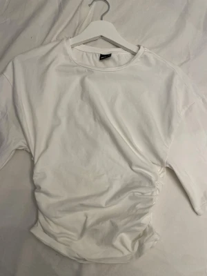 Vit basic t-shirt från Gina Tricot - En klassisk vit t-shirt från Gina Tricot i storlek XS. T-shirten har rund halsringning, korta ärmar och scrunch i sidorna. Tillverkad i mjuk bomull som känns skön mot huden. Perfekt att matcha med jeans eller kjol för en enkel och clean look.