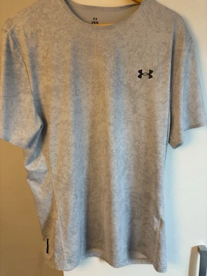 Ljusgrå träningströja från Under Armour rush - Snygg ljusgrå t-shirt från Under Armour med diskret mönster och svart logga på bröstet. Tillverkad i funktionsmaterial som andas, perfekt för träning. RUSH-kollektionen ger extra komfort och rörelsefrihet. Rund hals och korta ärmar.
