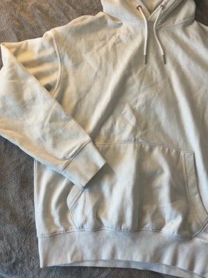 Vit hoodie med ficka framtill - Säljer en stilren vit hoodie med huva och dragsko. Hoodien har en stor magficka och ribbade muddar vid ärmslut och nederkant. Perfekt för en avslappnad look och enkel att matcha med andra plagg. Nopprig med små fläckar på ena arm, se bilder. 