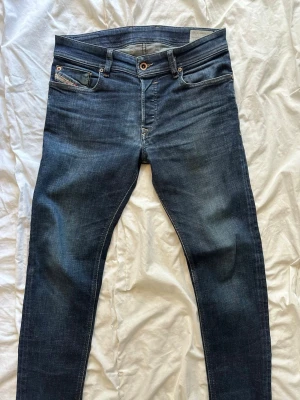 Mörkblå jeans från Diesel - Snygga mörkblå slim fit jeans från Diesel med klassisk femficksdesign och diskreta slitningar. Jeansen har en låg midja och smal passform som sitter tajt längs benen. Materialet är mjukt denim med stretch för extra komfort. 30:32