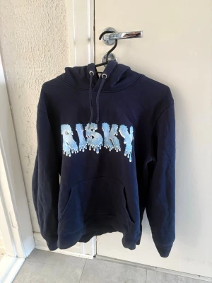 Mörkblå hoodie med RISKY-tryck - Mörkblå hoodie med stor ficka framtill och huva med dragsko. På bröstet står det 'RISKY' i stora, blåvita bokstäver med droppande effekt. Perfekt för en chill och avslappnad stil.