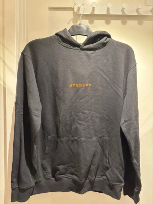 Svart hoodie från Mennace | S - Svart hoodie från Mennace med orange broderad logga på bröstet. Klassisk modell med huva och stor känguruficka framtill. Mjuk bomullsblandning som är skön att ha på sig. Perfekt för en chill och avslappnad stil.