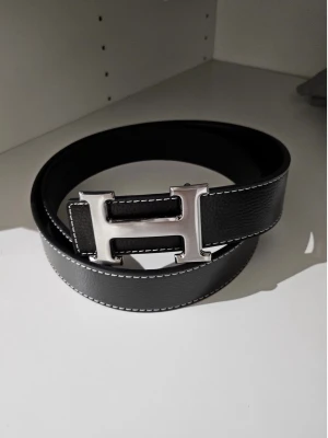 Svart H-spänne - Snyggt svart skärp med ett stort, silverfärgat H-spänne. Skärpet är gjort i skinn och har vita kontrastsömmar längs kanterna. Perfekt accessoar för att lyfta din outfit med en lyxig touch. Hör av dig vid intresse