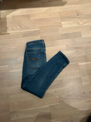 Nudie jeans - Säljer ett par klassiska blå jeans från Nudie Jeans Co med raka ben och snygga kontrastsömmar. Jeansen har en bakficka med Nudie-logga och är tillverkade i slitstark denim. Perfekta för en avslappnad och trendig look.