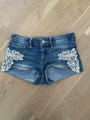 Blå jeansshorts med spets från H&M - Snygga blå jeansshorts från H&M med vita spetsdetaljer på sidorna. Klassisk femficksmodell med knapp och dragkedja framtill. Perfekta för varma dagar och ger en cool touch till din outfit.