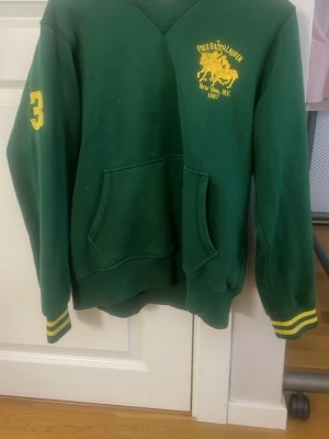 Grön hoodie från Polo Ralph Lauren - Grön hoodie från Polo Ralph Lauren med gul logga och hästtryck på bröstet, samt siffran 3 på ärmen. Har känguruficka framtill och ribbade muddar med gula ränder. Tillverkad i mjukt bomullsmaterial och har huva baktill. Koster över 1000 lappen egentligen