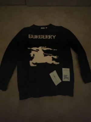 Svart stickad Burberry-tröja - Säljer en svart stickad tröja från Burberry med stort beige motiv av riddare och Burberry-logga framtill. Tröjan har rund hals och långa ärmar. Perfekt för dig som vill ha en statement-piece från ett klassiskt märke.