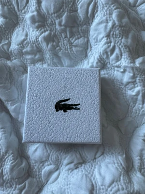 Svart armband från Lacoste - Snyggt svart armband från Lacoste i metall med länkdesign och diskret logga. Kommer i original ask med krokodilemblem på locket. Perfekt accessoar för dig som gillar stilrena detaljer och vill ha något som sticker ut lite extra.