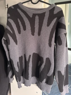 Grå stickad tröja med svart mönster - Säljer en oversized stickad tröja i grått med abstrakt svart mönster. Tröjan har rund halsringning, långa ärmar och ribbade muddar vid ärmslut och nederkant. Perfekt för dig som gillar streetwear och unik design. Helt ny