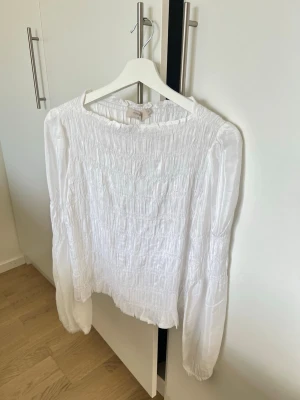 Vit smockad blus från Cream - Trendig vit blus från Cream med smockad överdel och långa ärmar. Blusen har en rak passform, rund halsringning och är gjord i ett lätt, luftigt tyg som ger en romantisk känsla. Perfekt att styla med jeans eller kjol.