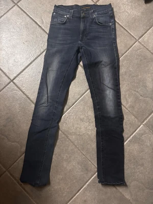 Mörkblå slim fit jeans från Nudie - Säljer ett par mörkblå slim fit jeans från Nudie Jeans med klassisk femficksmodell och snygga slitningar framtill. Jeansen har en rak passform och detaljerade sömmar på bakfickorna. Tillverkade i bomull med stretch för extra komfort. W27 L32