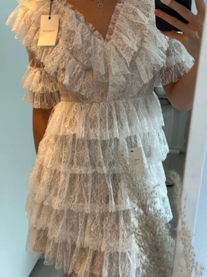 By Malina klänning - Superfin kort vit klänning från By Malina med flera lager volanger och v-ringning. Klänningen är gjord i spets och har korta ärmar. Perfekt för dig som vill sticka ut med en snygg och trendig look.