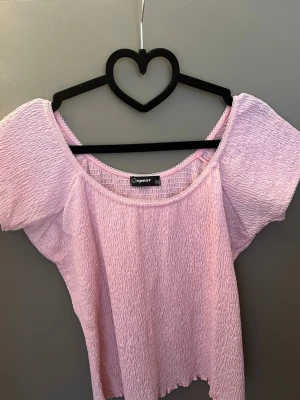 Ljusrosa smockad topp från Peppar - Supersöt ljusrosa topp från Peppar med smockad struktur och korta ärmar. Toppen har en vid, rund ringning och är lätt croppad med vågig kant nedtill. Perfekt att styla med jeans eller kjol för en trendig look.