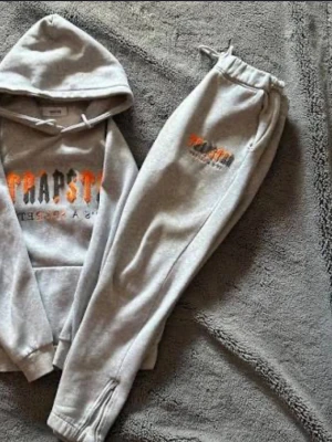 Grå hoodie och joggers från Trapstar - Säljer ett set med grå hoodie och matchande joggers från Trapstar. Hoodien har stor huva, magficka och coolt tryck i orange och svart på bröstet. Joggers har samma tryck på benet och dragsko i midjan. Perfekt för chill dagar och streetstyle. Materialet är mjukt och bekvämt.