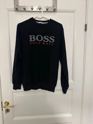 Svart sweatshirt från Hugo Boss - Svart sweatshirt från Hugo Boss med stort BOSS-tryck och broderad HUGO BOSS-text i rött på bröstet. Tröjan har rund halsringning och ribbade muddar vid ärmslut och nederkant. Perfekt för en stilren och avslappnad look. Storlek M