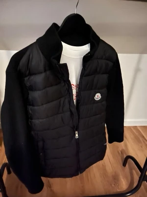 Svart pufferjacka från Moncler - Snygg svart pufferjacka från Moncler med quiltad front och stickade ärmar. Jackan har dragkedja framtill, hög krage och Moncler-logga på bröstet. Perfekt för kalla vinterdagar och ger en clean, modern look.