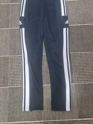 Svarta Adidas träningsbyxor - Säljer ett par svarta träningsbyxor från Adidas med klassiska vita ränder längs benen och Adidas-logga på låret. Byxorna har smal passform och är tillverkade i ett mjukt och stretchigt material som passar perfekt till träning eller chill. Det står L på de men skulle säga att de passar S / m mer.