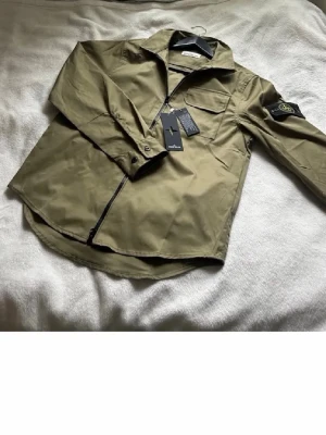 Olivgrön overshirt från Stone Island - Snygg olivgrön overshirt från Stone Island med klassisk patch på ärmen och hel dragkedja framtill. Overshirten har krage, bröstficka och långa ärmar med knapp vid manschetten. Perfekt för lager på lager och streetwear-stil.