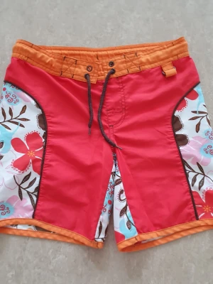 Badshorts - Säljer ett par färgglada badshorts med röd front, orange midja och benkant samt blommigt mönster i vitt, rosa, blått och brunt på sidorna. Snörning i midjan och coola kontrastdetaljer. Perfekta för stranden eller poolen!