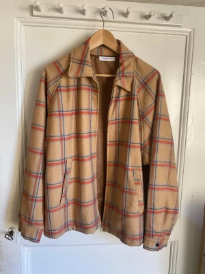 Beige rutig jacka från Uniqlo Lemaire - Snygg beige jacka från Uniqlo Lemaire i storlek L med rutigt mönster i rött och blått. Jackan har dragkedja, två fickor fram och klassisk krage. Materialmix av ull, polyester, akryl, nylon och viskos. Perfekt för dig som gillar retrovibbar och stilren design.