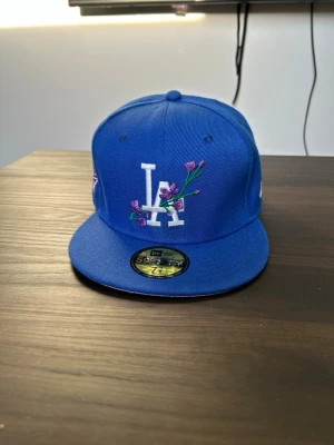 Blå LA New Era keps med blommor - Säljer en blå New Era 59FIFTY keps med LA-logga framtill och broderade lila blommor. Klassisk flat brim och broderad New Era-logga på sidan. Perfekt för dig som vill sticka ut med en unik och snygg keps.