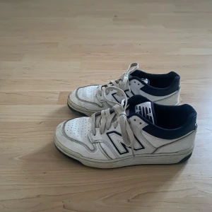 Vita New Balance sneakers med blå detaljer - Säljer ett par vita New Balance sneakers med mörkblå detaljer och svart sula. Klassisk låg modell med perforerad tå, stora N-loggor på sidorna och bekväm vadderad kant. Skorna har snörning och är tillverkade i läder och textil. 